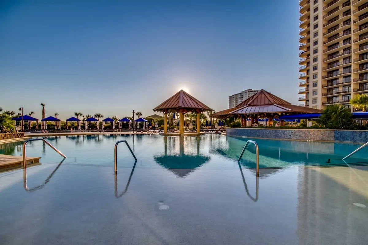 This exterior image features a tranquil swimming pool surrounded by tropical cabanas and a high-rise building, all set against a dramatic sunset sky. The setting hints at evening leisure, sunset cocktails, and luxurious amenities, aligning with the idea of winding down after a day of spa treatments or exploring local attractions.