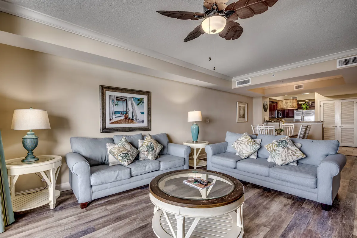 A welcoming living area with plush sofas, a central coffee table, a floor lamp, wall art, and a ceiling fan. The scene radiates comfort and is ideal for relaxation or social gatherings.
