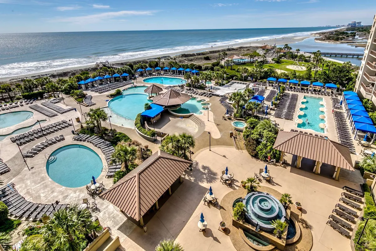 This exterior image captures a beautifully landscaped pool area with cabana structures, inviting lounge chairs, and a stunning ocean view in the background. The tranquil water and thoughtfully arranged gardens create a sense of exclusive relaxation, ideal for couples seeking a romantic escape. The scene suggests both private moments and leisurely afternoons by the pool, with the ocean just steps away.