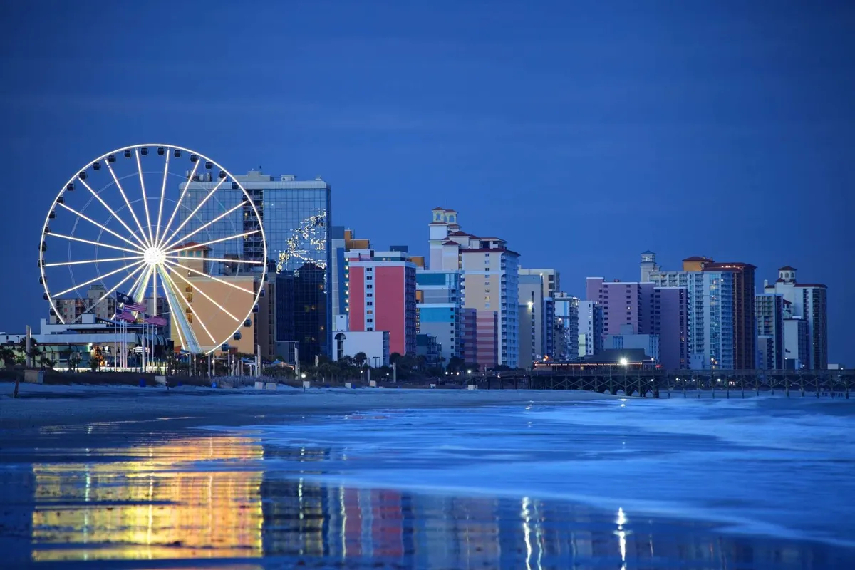 A vibrant exterior shot featuring a brightly lit Ferris wheel, colorful buildings, and their reflections dancing on the water. The festive mood signals lively nightlife and entertainment nearby.