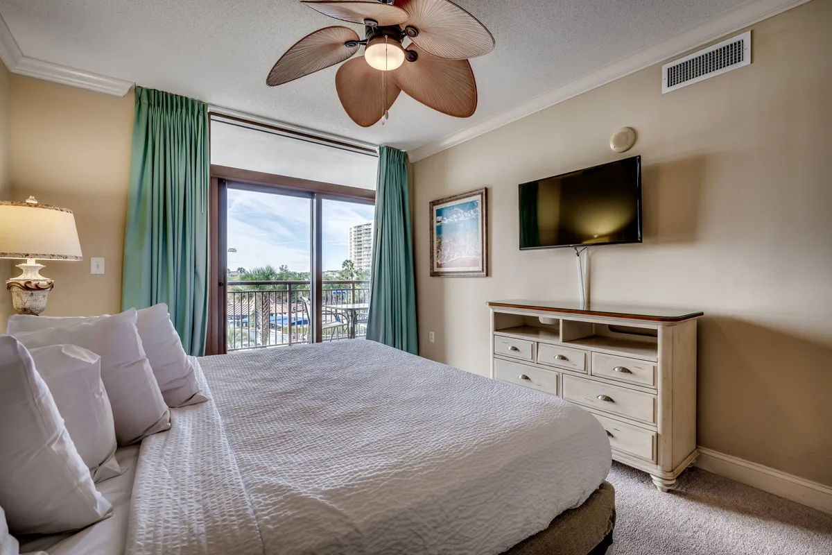 A serene bedroom featuring a king-sized bed draped in plush linens, a large window letting in natural light, a ceiling fan overhead, a wall-mounted TV, and a dresser. The calming palette and uncluttered layout evoke a restful atmosphere.