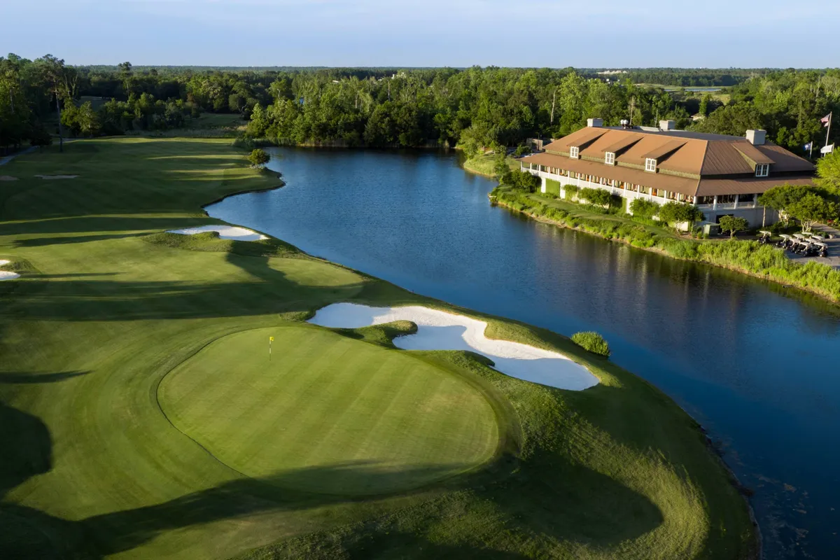 A lush golf course stretches beside a sparkling lake, with sand traps leading up to a welcoming clubhouse. The setting is serene and sunlit, inviting guests to enjoy golf, nature walks, or social events.