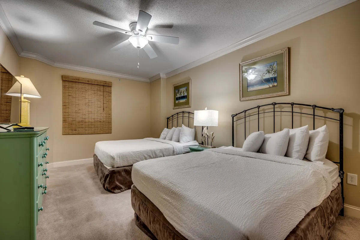 A guest bedroom with two double (queen-sized) beds, a green dresser, warm bedside lighting, and framed artwork. The space feels cozy and inviting, perfect for families or groups.