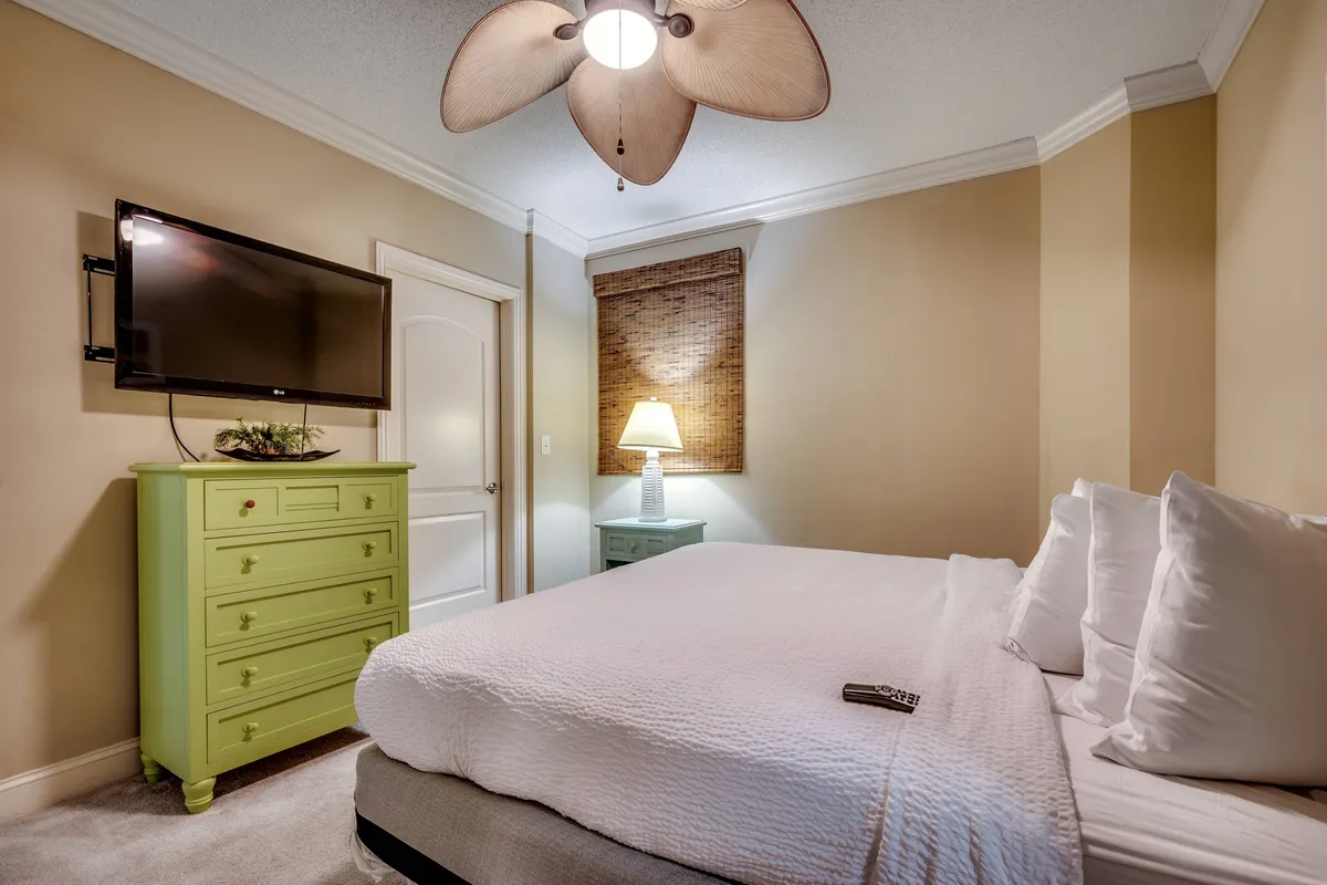 A guest bedroom featuring a bed with crisp white linens, a green dresser, a TV on the wall, and a softly glowing lamp. The setting feels inviting and private, ideal for unwinding.