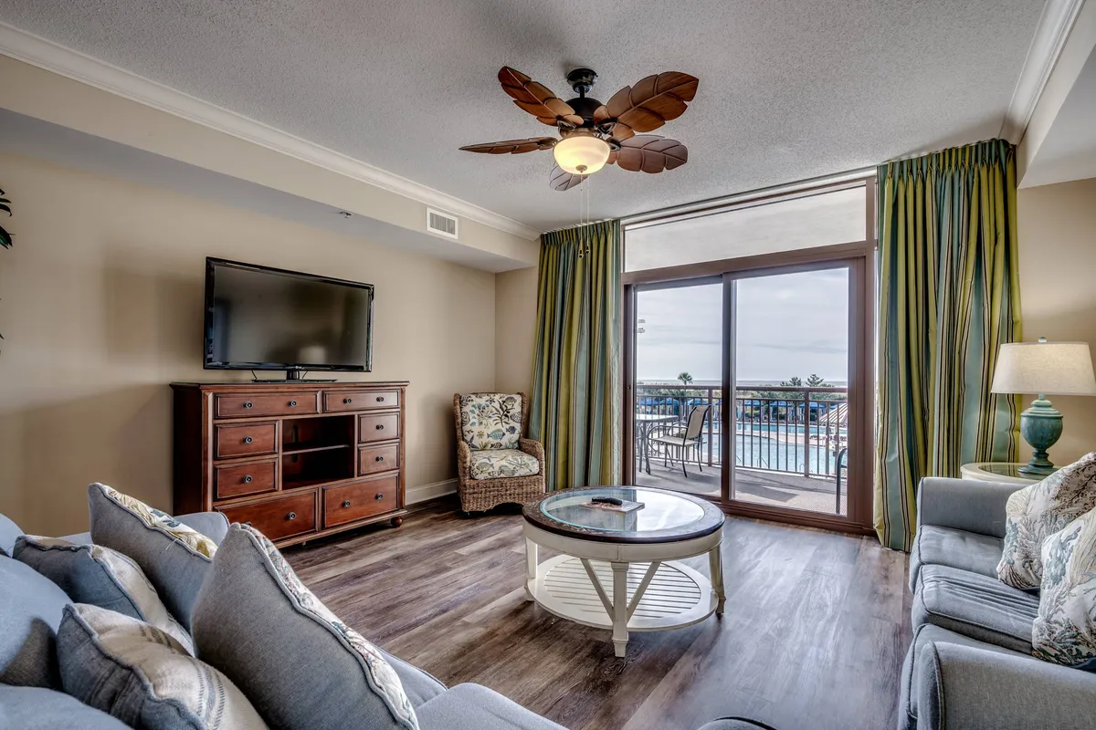 The image showcases a stylish, sunlit living room with a plush sofa and coffee table arranged for conversation, a flat-screen TV for entertainment, and large windows offering a glimpse of the balcony and the sunset beyond.